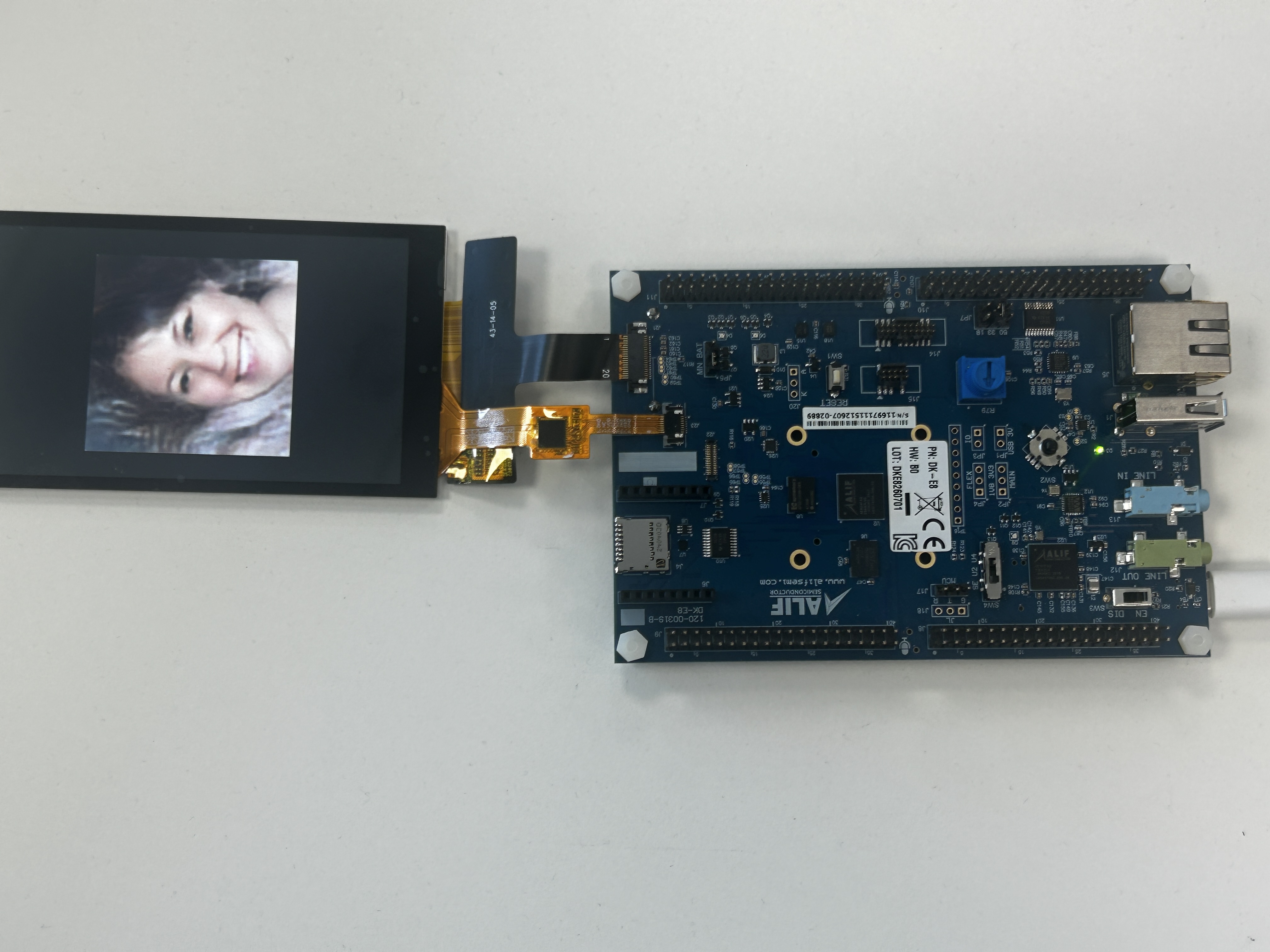Image Alt Text:Photograph of the Alif Ensemble E8 DevKit with its attached LCD displaying a generated smiling face. The display confirms that the pico-faces application completed inference and rendered the 128 x 128 result on the board.