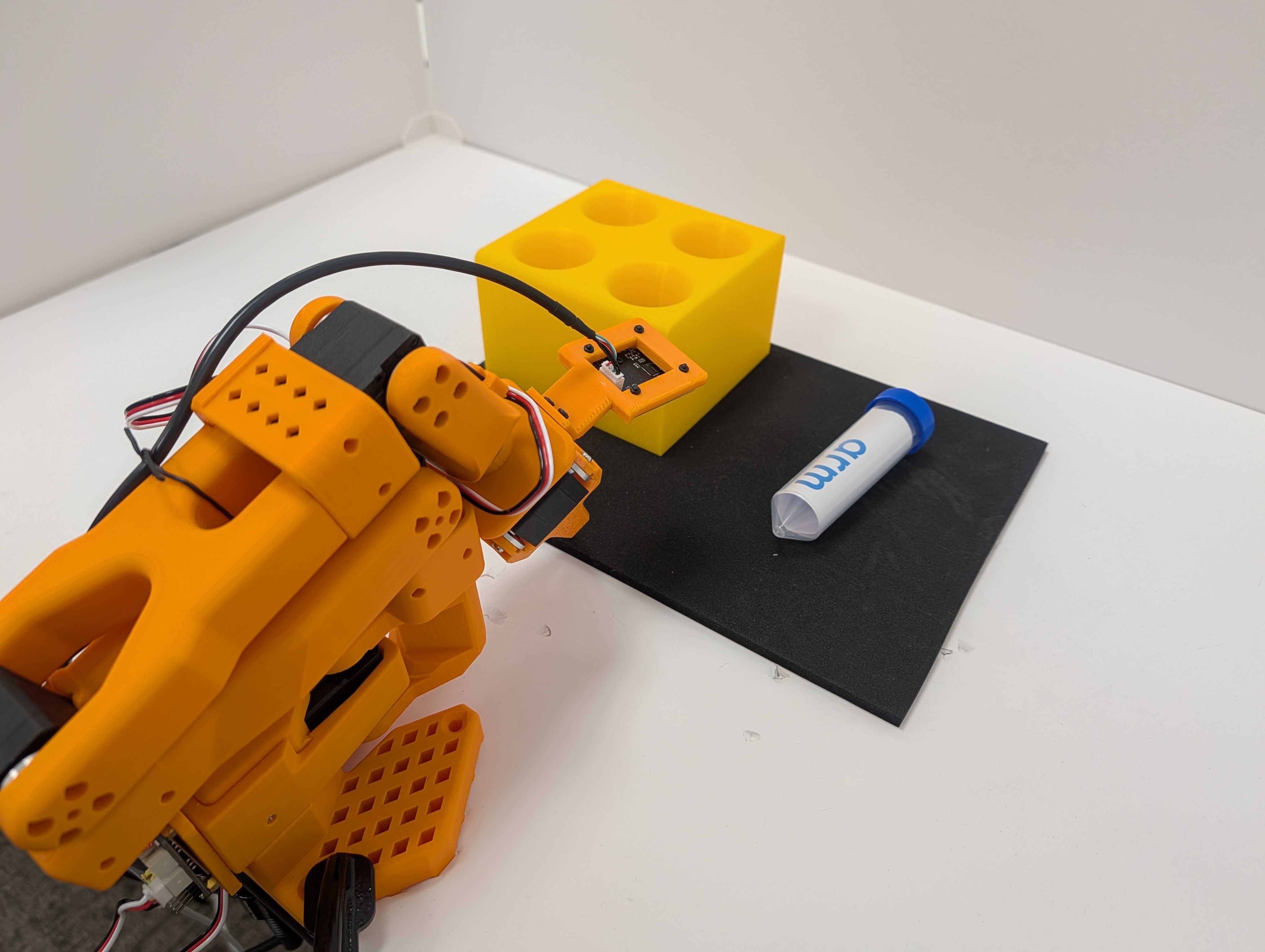 Image Alt Text:SO-101 robot arm beside a vial and four-hole yellow rack. The black task mat keeps the pickup and placement area visually distinct for data collection.