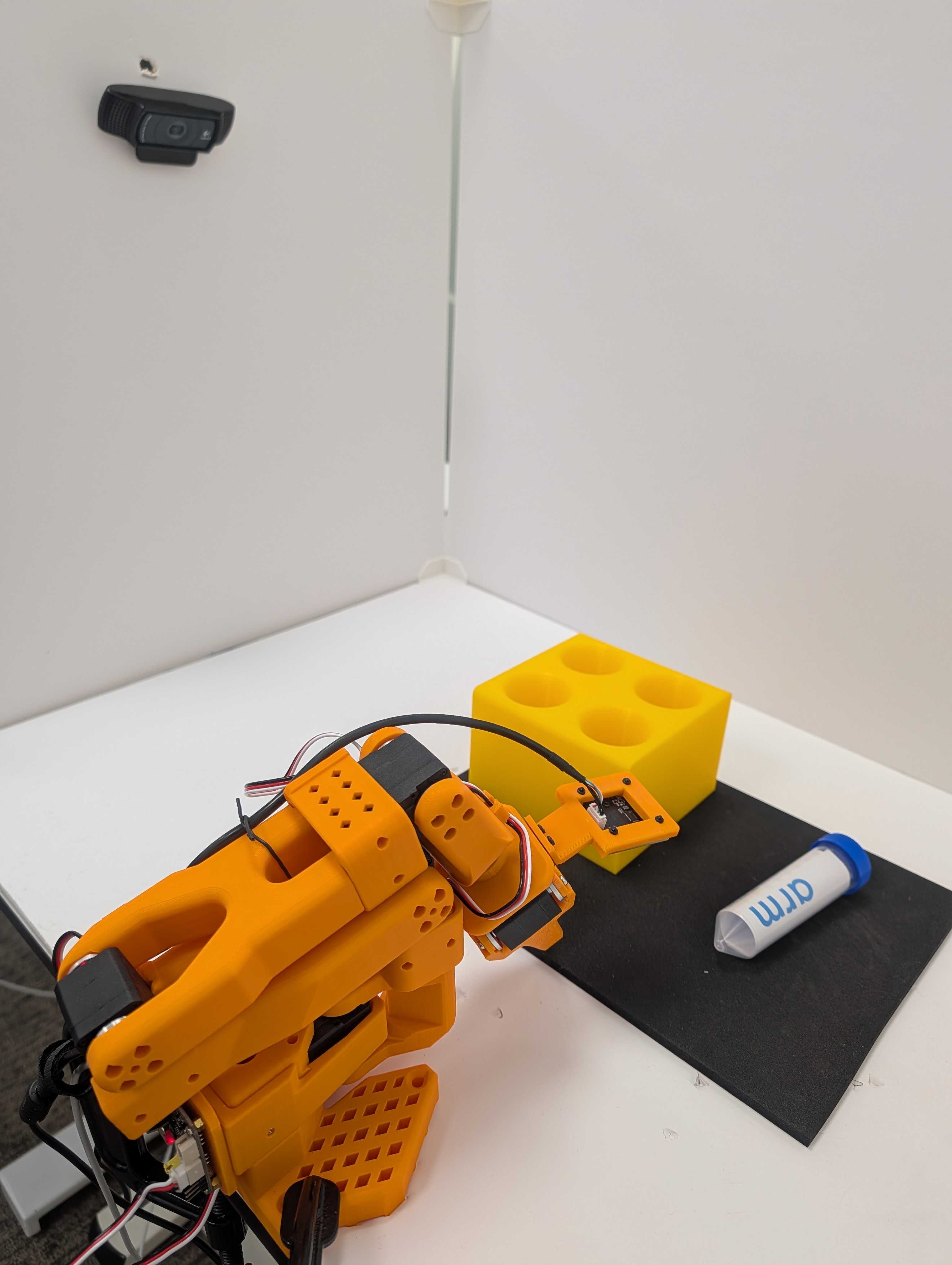 Image Alt Text:SO-101 follower workspace with one camera mounted near the gripper and a second camera fixed above the table. The fixed camera covers the vial, yellow rack, follower, and motion area while the gripper camera provides the close manipulation view.